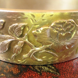 May include: A gold-toned metal bangle bracelet with an embossed floral design. The bracelet features a detailed rose and leaf pattern, with silver-toned accents. The design is raised, creating a three-dimensional effect. The bracelet has a smooth, polished finish.