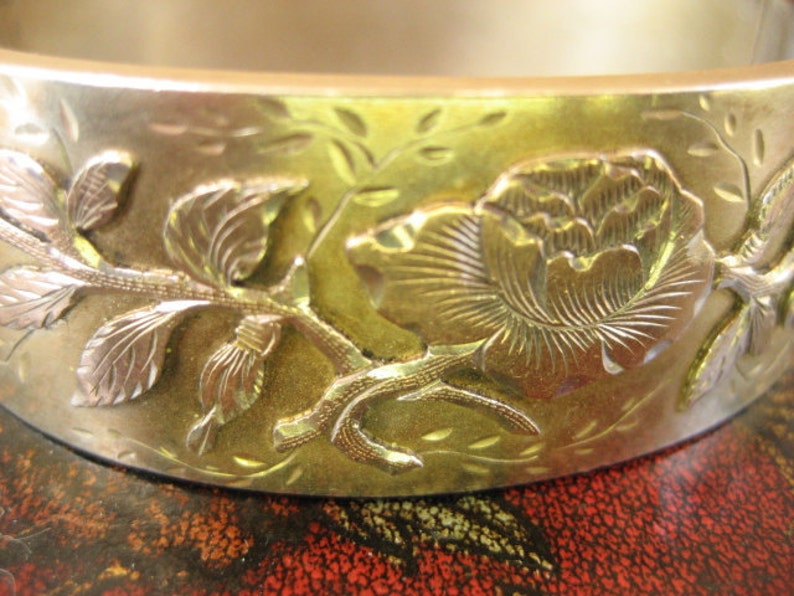 May include: A gold-toned metal bangle bracelet with an embossed floral design. The bracelet features a detailed rose and leaf pattern, with silver-toned accents. The design is raised, creating a three-dimensional effect. The bracelet has a smooth, polished finish.