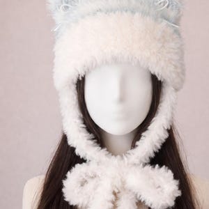 May include: A fluffy, faux-fur hat featuring a light blue and white color scheme with two rounded ear-like protrusions on top. The hat includes long, plush side panels that extend downward and tie together in a bow under the chin area.