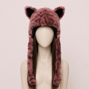 May include: A plush, faux fur hat in a dusty rose and dark brown tiger-striped pattern. The hat features two upright, pointed ears with black inner lining and two long, dangling side flaps that extend downward from the main cap structure.
