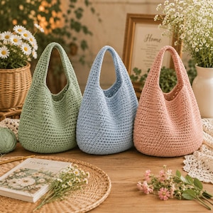 May include: Three handmade crochet bags in soft pastel colors: sage green, light blue, and dusty pink. Each bag features a rounded, slouchy silhouette with a single integrated handle, crafted from a textured yarn in a consistent stitch pattern.