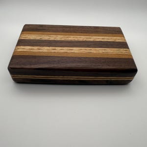May include: A rectangular wooden box with a striped design. The box is made of various shades of brown wood, including dark brown, medium brown, and light brown. The stripes run horizontally across the top and sides, creating a visually appealing pattern.