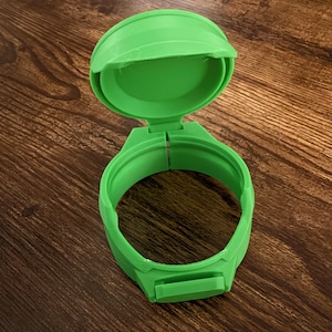 May include: A bright green, hinged, circular object, possibly a wearable accessory or container. The object is open, revealing an interior space. The design features a rounded top and a clasp. The item is made of plastic.