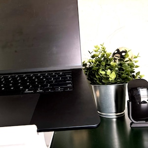 May include: A black laptop with a keyboard sits on a dark surface. A small potted plant with green leaves and a decorative skull is next to a black wireless mouse and charging stand. The background is a white wall.
