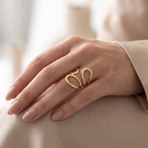 Könnte beinhalten: Ein goldfarbener Ring mit einem offenen, fließenden Design, das aus mehreren glatten, abgerundeten Schlaufen besteht, die sich um den Finger legen. Das Metall hat eine polierte, reflektierende Oberfläche und bildet eine durchgehende Form.