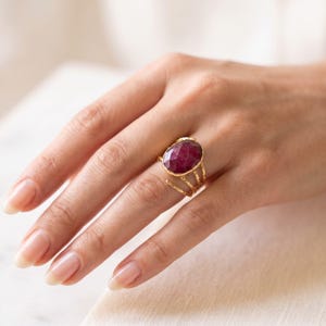 Könnte beinhalten: Ein Goldring mit einem großen, ovalen, facettierten, tiefroten Edelstein in einer Zargenfassung. Die Ringschiene besteht aus mehreren dünnen, strukturierten Golddrähten, die zusammenlaufen, um die zentrale Steinfassung zu stützen.