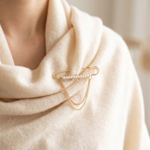 Può includere: Una spilla da balia color oro con una fila di piccole perle bianche e cristalli trasparenti lungo la barra, dotata di due delicate catenelle color oro che pendono in un anello drappeggiato dalle estremità, fissata su un morbido tessuto color crema.