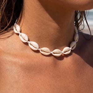 May include: A choker necklace featuring a series of off-white cowrie shells strung together on a thin white cord. The shells are arranged horizontally, creating a continuous band that rests closely around the neck, showcasing their natural texture.