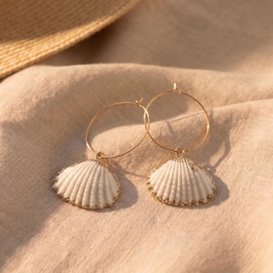 May include: A pair of gold-toned hoop earrings, each featuring a single white, ribbed seashell charm suspended from the bottom. The shells have a natural, fan-like shape with distinct vertical ridges, and the hoops are thin, circular metal wires.