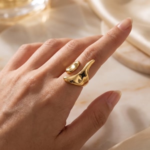 Op de afbeelding: Een goudkleurige ring met een open, asymmetrisch ontwerp met twee afzonderlijke, gladde, organisch gevormde elementen die om de vinger wikkelen. Het oppervlak van het metaal is gepolijst en reflecterend, wat een vloeiend, vloeibaar uiterlijk creëert.