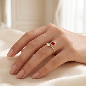 Puede incluir: Un anillo de oro con una pequeña piedra preciosa redonda y transparente y una piedra roja más grande en forma de lágrima. La banda es delgada y está decorada con pequeñas piedras transparentes a lo largo del lado, creando un aspecto refinado.