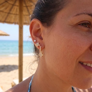 May include: A set of gold-toned earrings featuring a small stud with a clear stone, a fish-shaped stud with baguette-cut stones, and a hoop earring with a dangling fish charm that also features baguette-cut stones and a small red accent stone.