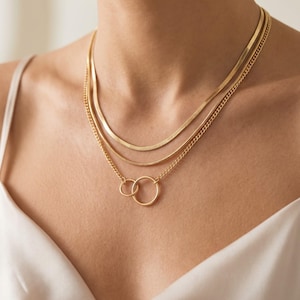 Può includere: Un set di collane a strati color oro composto da tre catene distinte. Lo strato superiore è una catena a spina di pesce piatta, quello centrale è una sottile catena a serpente e lo strato inferiore presenta una delicata catena a maglie con due anelli circolari intrecciati.