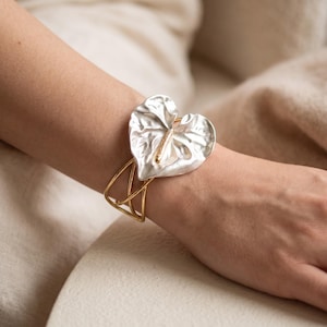 Può includere: Un bracciale a fascia con un grande ornamento a forma di cuore a foglia di anthurium color argento, caratterizzato da una superficie testurizzata e uno spadice centrale. Il bracciale è composto da sottili fili metallici dorati intrecciati.