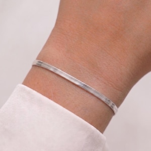 Könnte beinhalten: Ein dünnes, silberfarbenes Fischgräten-Kettenarmband mit flachem, flexiblem Design. Das Armband weist ein eng gewebtes, pfeilförmiges Muster auf, das eine glatte, reflektierende Oberfläche erzeugt. Es wird am Handgelenk getragen und hat ein metallisches Finish.