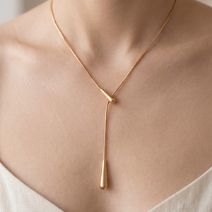 May include: A gold-toned lariat necklace featuring a thin snake chain and two smooth, teardrop-shaped pendants. One pendant is fixed at the intersection point, while the other hangs at the end of the chain, creating a Y-shaped silhouette.