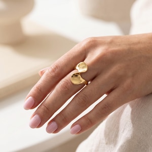 May include: A gold ring featuring a smooth, polished surface with a distinct wave-like, undulating shape that wraps around the finger. The metal has a reflective finish, highlighting the curved, organic contours of the band design.