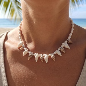 May include: A necklace composed of small, spiraled seashells in shades of white, cream, and light brown. The shells are strung together in a continuous row, creating a textured, organic design that rests around the neck area.