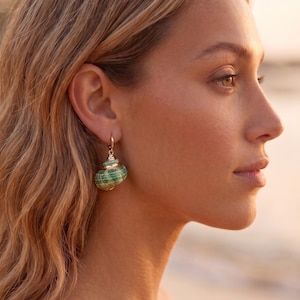 Peut inclure: Une boucle d'oreille dorée composée d'une petite breloque en forme de coquillage en spirale, ornée de rayures horizontales vertes et blanches. Le coquillage est suspendu à un anneau métallique doré simple fixé au lobe de l'oreille.