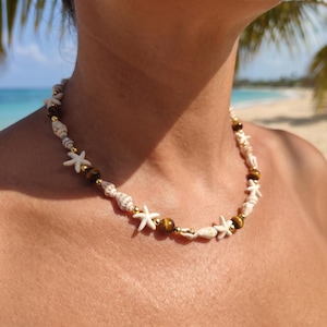 May include: A beaded necklace featuring white seashell charms, small white starfish accents, brown tiger eye stone beads, and small gold-toned metallic spacer beads arranged in a repeating pattern around the entire length of the circular strand.