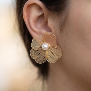 May include: A gold-toned earring shaped like a flower with delicate, thin wire petals arranged in a layered pattern. At the centre of the floral design sits a single, smooth, round white pearl, providing a contrast to the metallic gold wire structure.