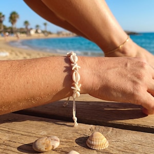 Peut inclure: Un bracelet composé d'une série de petits coquillages et de formes d'étoiles de mer blanc cassé enfilés sur un cordon clair. Le bracelet est réglable avec des extrémités de cordon pendantes et est porté au poignet, affichant un style naturel.