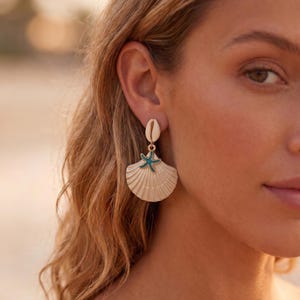 Peut inclure: Une boucle d'oreille pendante composée d'un petit coquillage cauri blanc en haut, relié à un coquillage Saint-Jacques en éventail de couleur crème. Une petite breloque en forme d'étoile de mer bleu sarcelle est fixée au centre.