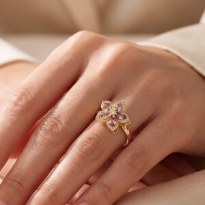 May include: A gold ring featuring a flower-shaped centerpiece with five marquise-cut light purple stones arranged as petals. The petals are outlined with small clear stones, and a single clear stone is set in the center of the flower design.