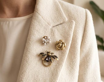 May include: Three decorative pins attached to a cream-colored textured blazer. One pin is a gold-toned bee with a dark central stone, one is a gold-toned flower, and one is a circular cluster of small white pearls surrounding a clear center stone.