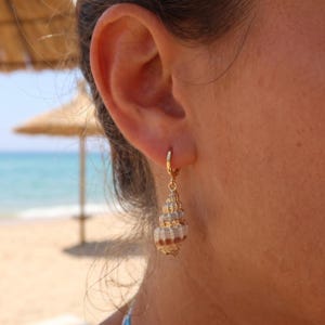 May include: A gold hoop earring featuring a small, natural spiral seashell charm. The seashell has a textured, conical shape with white and light brown horizontal bands. The earring is attached to the earlobe, showcasing the detailed shell structure.