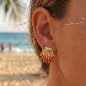 Può includere: Un orecchino a lobo dorato a forma di conchiglia di capasanta, caratterizzato da un design sfumato che passa da un colore crema pallido nella parte superiore a una tonalità rosso-arancio vibrante e satura sul bordo inferiore, con striature.