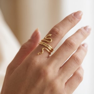 Puede incluir: Un anillo dorado con forma de serpiente enroscada que se envuelve alrededor del dedo. La banda de metal presenta un acabado liso y pulido con un diseño definido de cabeza y cola, creando un efecto de espiral continua alrededor del dedo.
