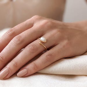 Puede incluir: Un anillo abierto de tono dorado que presenta una perla blanca de forma ovalada en un extremo y una pequeña esfera dorada pulida en el otro. La banda es delgada y lisa, diseñada para rodear parcialmente el dedo con una abertura ajustable.