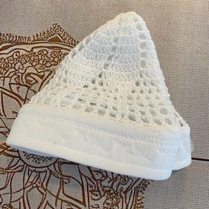 May include: A white crocheted skullcap with a solid white band. The skullcap features an open, airy design, and the band is made of a smooth, solid fabric. The cap is likely handmade and rests on a patterned surface.