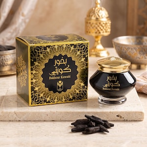 Op de afbeelding: Een zwart-gouden vierkante doos en een bijpassende zwarte glazen pot met een gouden deksel, beide gelabeld Bakhour Kuwaiti in Arabisch en Engels schrift. Een kleine hoop donkere, houtachtige wierookchips ligt voor de doos en de pot.