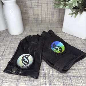 May include: Black fingerless gloves with circular, iridescent emblems. One glove is leather, the other is a stretchy fabric. The emblems feature a white and black design on one glove and a colourful design on the other.