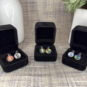 May include: Three pairs of earrings in black velvet boxes. Each pair features a round pendant with colorful designs. The earrings are displayed on a patterned surface, with white vases and greenery in the background.