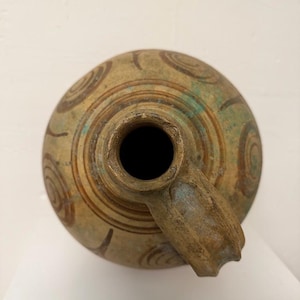 May include: Antique ceramic jug with a spout, featuring a circular body adorned with brown spiral patterns. The jug has a weathered, aged appearance with hints of green and brown hues, suggesting its age and use.