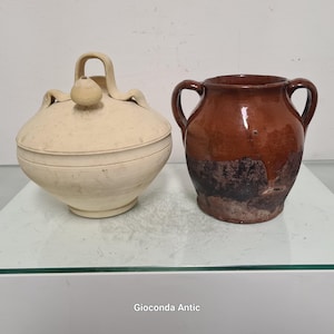Puede incluir: Dos vasijas de cerámica antiguas. Una es un tarro con tapa de color crema con asa y pomo en la parte superior. La otra es una olla marrón con dos asas y una base más oscura. Ambas se exhiben sobre una superficie de vidrio.