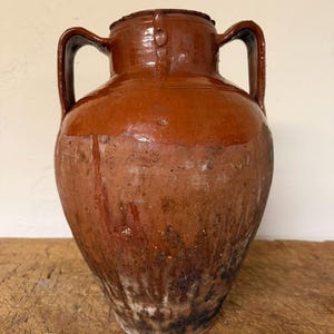 Puede incluir: Una vasija de terracota grande y bulbosa con dos asas laterales y un cuello estrecho. La parte superior está recubierta con un esmalte marrón liso y brillante, mientras que el cuerpo inferior muestra una superficie de arcilla roja, rugosa y texturizada.