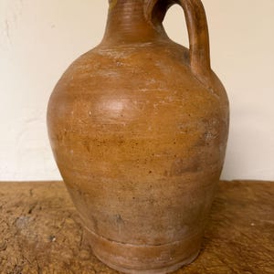 Puede incluir: Una jarra de barro marrón de aspecto rústico con cuerpo redondeado, cuello estrecho y una sola asa curva en el lateral. La superficie presenta una textura mate y desgastada con sutiles estrías horizontales y signos visibles de antigüedad.