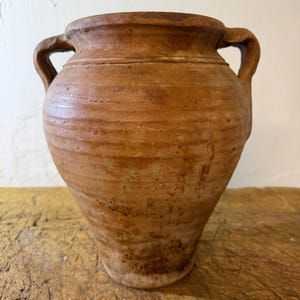 May include: A rustic, medium-sized terracotta vessel with a wide, circular opening and two symmetrical side handles. The exterior surface features a textured, horizontal ribbed pattern with natural color variations in shades of brown and tan.