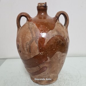 Puede incluir: Una jarra de cerámica antigua marrón con dos asas y un cuello estrecho. La jarra tiene un cuerpo redondeado con un esmalte marrón oscuro y parches marrones claros. La jarra es probablemente una pieza antigua o vintage.