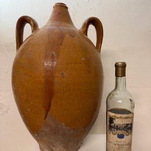 Puede incluir: Una jarra grande de cerámica marrón antigua con dos asas, junto a una botella de vino de vidrio transparente con una etiqueta. La jarra tiene una base ancha y un cuello estrecho. La botella de vino tiene una tapa dorada y una etiqueta con texto.