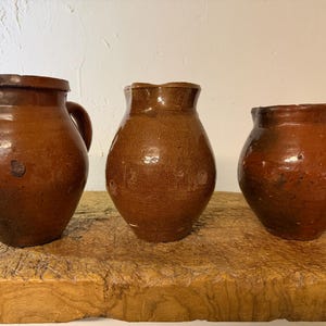 Puede incluir: Tres jarras de cerámica marrón con un acabado brillante y esmaltado. Cada jarra presenta un cuerpo redondeado, una boca ancha y un asa resistente. Los recipientes muestran variaciones en color y textura, exhibiendo una apariencia rústica y terrosa con imperfecciones superficiales visibles y un tono marrón cálido y profundo en todo el conjunto.