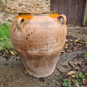 May include: A large, antique terracotta pot with a wide, ribbed body and two curved handles. The pot has a weathered, aged appearance with a faded orange and brown color. The rim is a darker orange. The pot is outdoors on a concrete surface.