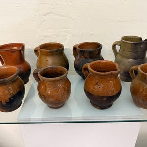Puede incluir: Colección de ocho jarras de cerámica rústicas hechas a mano. Las jarras varían en tamaño y color, con tonos marrones, naranjas y negros. Cada jarra tiene un asa y un pico vertedor.
