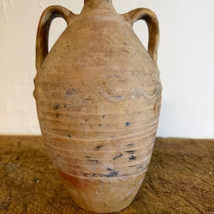 May include: A tan, weathered earthenware jug with two side handles and a narrow neck. The body features horizontal incised lines and a wavy pattern around the upper section, with visible dark scuffs and reddish-brown patches on the surface.