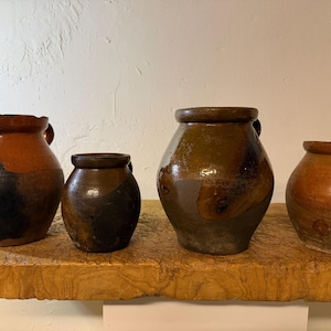 Puede incluir: Una colección de cuatro vasijas de cerámica rústica con cuerpos redondeados y cuellos cortos y anchos. Los recipientes presentan tonos tierra de marrón, bronceado y carbón oscuro con un acabado de esmalte brillante e irregular.