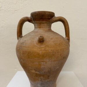May include: A tan-colored, antique ceramic jug with two handles and a small spout. The jug has a rounded body and a slightly flared base. The surface shows signs of age and wear, with variations in color and texture. The jug is displayed on a white surface.